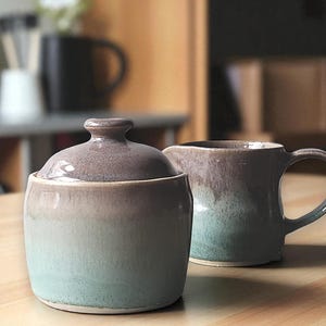 May include: A ceramic sugar bowl with a lid and a creamer. The set is glazed in a gradient of colours, transitioning from a muted purple at the top to a soft teal at the base. The pieces are on a wooden surface.