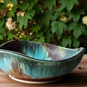 May include: A handmade ceramic bowl with an unusual, organic form. The bowl has a mix of turquoise, brown, and green glaze, producing a marbled appearance. The bowl is placed on a wooden surface, with a blurred green backdrop.