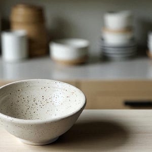 May include: A speckled, off-white ceramic bowl with a rounded shape. The bowl has a slightly angled rim and sits on a wooden surface. Other ceramic pieces are visible in the background, suggesting a kitchen or dining setting.