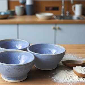 May include: Three handmade ceramic bowls with a light blue glaze. The bowls are on a wooden surface with flour and wooden spoons. The bowls have a rounded shape and a white base. Kitchenware.