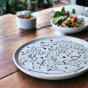 May include: A white ceramic plate with a black abstract design. The design features wavy lines, dots, and swirls.