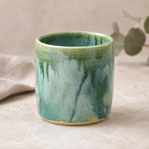 May include: A cylindrical ceramic mug featuring a reactive glaze with blended shades of teal, sage green, and deep forest green. The surface displays a smooth, glossy finish with vertical drip patterns cascading from the rim towards the base.