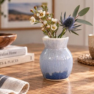May include: A small, bulbous ceramic vase with a glossy glaze that transitions from a pale, icy blue at the neck to a deep, saturated navy blue at the base. The vase holds a small bouquet of white flowers and a single spiky blue thistle stem.