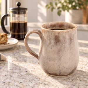 May include: A ceramic mug with a rounded body and a curved handle, featuring a mottled, reactive glaze in shades of cream, beige, and dark brown. The mug has a wide rim and a smooth, textured finish, showcasing a blend of earthy, neutral tones.