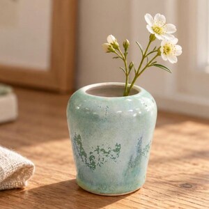 May include: A small, rounded ceramic vase with a light turquoise and seafoam green mottled glaze. The vase features a smooth, wide opening and contains a single delicate stem with three small white flowers with yellow centres.