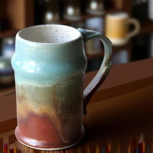 May include: A handcrafted ceramic mug with a distinctive, textured design. The mug displays a gradient of colours, including blue, green, and red-brown. The handle is a darker shade of brown. This stoneware mug is ideal for coffee or tea.