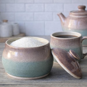 May include: A ceramic tea set featuring a sugar bowl filled with white granules, a mug with dark liquid, and a teapot. The set has a two-tone design, with a brown top and a teal bottom. The sugar bowl's lid is off to the side.