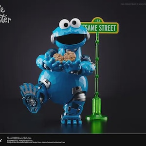 以下が含まれることがあります： ファセット加工されたメタリック仕上げの青いクッキーモンスターのロボットフィギュアが、2枚の茶色のチョコチップクッキーを持っています。フィギュアの横には緑色のセサミストリートの標識があります。Cookie Monster、Blitzway、Carbotixの文字が見えます。