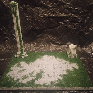 Op de afbeelding: Een miniatuur diorama scène met een stenen platform met een gebarsten tegelontwerp, omgeven door groen gebladerte. Twee witte zuilen met groene wijnranken zijn op het platform geplaatst, waardoor een klassieke esthetiek ontstaat.