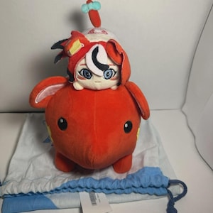 May include: A red plush toy with a smaller red plush toy on top. The smaller toy has a white face with blue eyes and a black crescent moon on its forehead. The larger toy has a yellow star on its side. The toys are sitting on a blue and white drawstring bag.