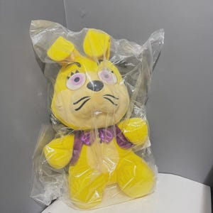 Puede incluir: Un juguete de peluche de conejo amarillo con orejas grandes, ojos de color violeta claro y nariz negra. El personaje lleva un chaleco morado con una pajarita morada a juego. El peluche está encerrado en una bolsa de plástico transparente.
