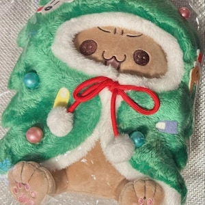 May include: A soft toy cat dressed as a Christmas tree. The green tree costume has white edging, colourful baubles, and a red bow. The cat has a light brown face and paws with pink paw prints.