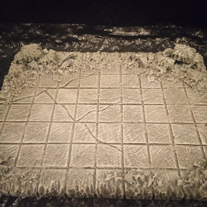 May include: A white, square, tiled gaming board with a cracked surface. The board is surrounded by a rough, textured border.