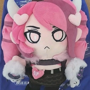 Op de afbeelding: Een pluche poppetje met roze en donkerpaars haar, witte hoorns en hartvormige accenten. De pop heeft grote ogen, een zwart topje, een zwarte rok met een roze hartdetail en witte harige manchetten aan de handen. Het personage heeft een kleine, eenvoudige mond en donkere haarpunten.