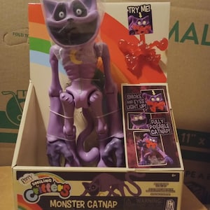 May include: A purple Monster Catnap action figure from the Smiling Critters series, packaged in a cardboard box with a rainbow graphic. The figure features a yellow crescent moon on its chest, large white eyes, and a red smoke effect accessory.