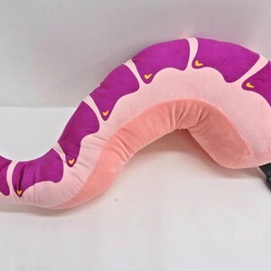 May include: A pink and purple plush worm-shaped pillow with a black tail. The pillow is designed to be used as a body pillow.