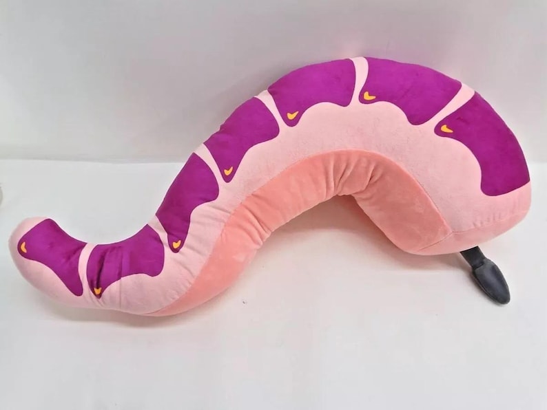 May include: A pink and purple plush worm-shaped pillow with a black tail. The pillow is designed to be used as a body pillow.