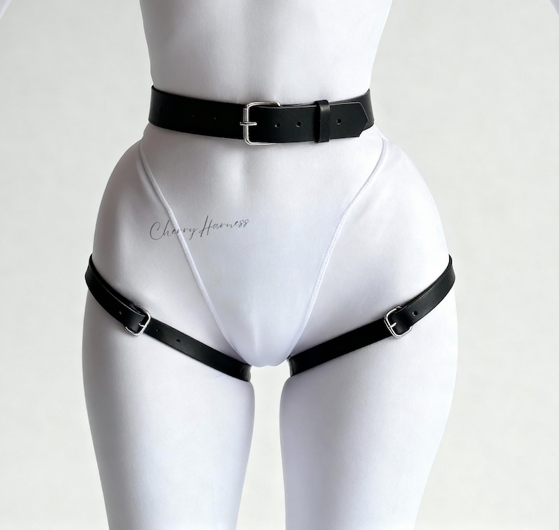 May include: Black leather body harness with silver-tone buckles. The harness includes a waist belt and leg straps. The text "Cherry Harness" is visible on the garment. The harness is designed to be worn over clothing.