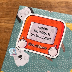 May include: A red and white Etch A Sketch toy with a white screen that says "YOUR NAME", "STREET ADDRESS", "CITY, STATE, ZIPCODE", and "Etch A Sketch".