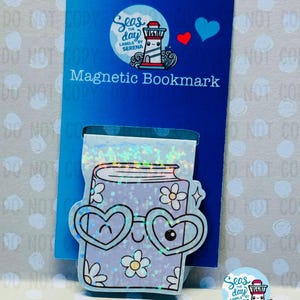 May include: A purple book-shaped magnetic bookmark with a glittery finish. The bookmark has heart-shaped sunglasses and a happy face. The bookmark is on a blue background with the text "Seas the day Labels by Serena" and "Magnetic Bookmark".