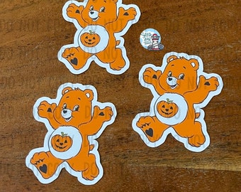 May include: Three orange and white cartoon bear stickers with black eyes and a black heart on each foot. Each bear is holding a jack-o-lantern pumpkin.