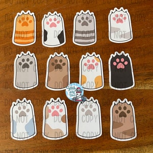 May include: Twelve paw print stickers with different patterns and colors. The paw prints are white with black outlines and feature different patterns like stripes, polka dots, and camouflage.