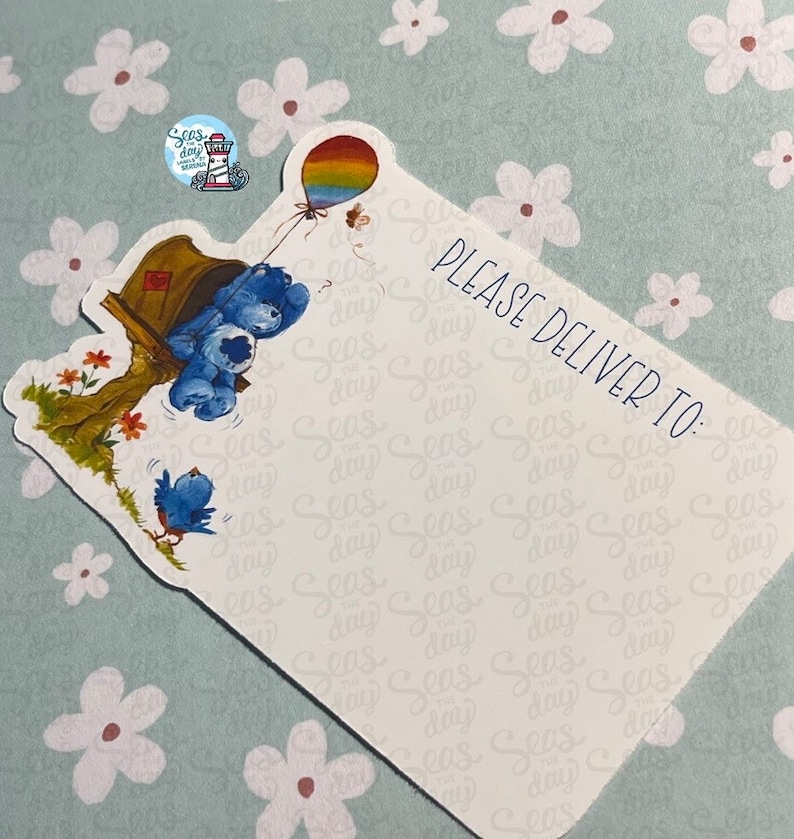 May include: A blue and white card with a cartoon illustration of two blue bears holding a rainbow balloon. The card has the text "PLEASE DELIVER TO:" printed on it.