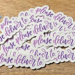 May include: A collection of white stickers with the phrase "please deliver to:" in a purple script font. The stickers are scattered on a light brown wooden surface. A small logo with a lighthouse is in the upper left corner.
