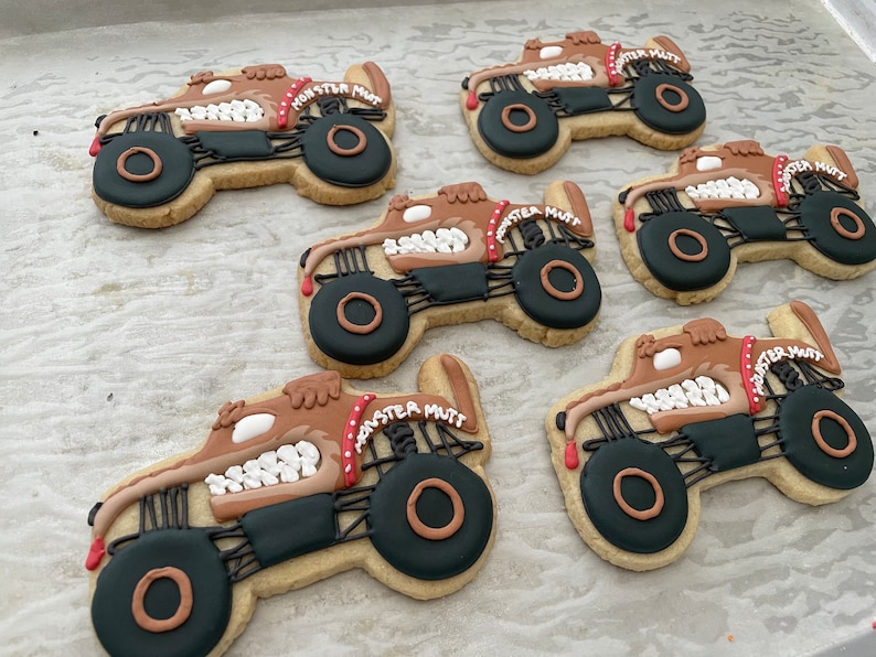 May include: Six decorated sugar cookies shaped like monster trucks. Each cookie is brown with black and white details. The cookies are decorated with the words "Monster Mutt" in red and white lettering.