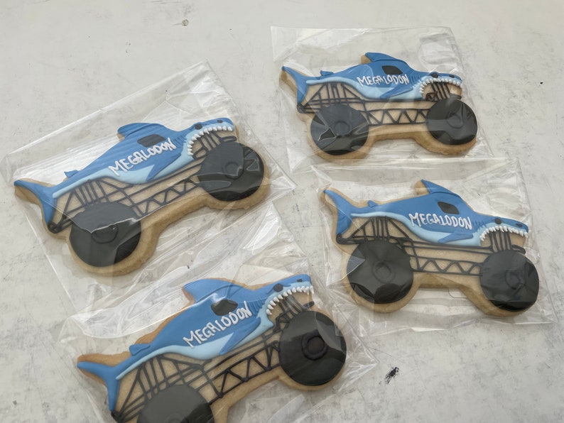 May include: Four blue and white decorated sugar cookies shaped like monster trucks with shark fins. Each cookie has the word "Megalodon" written on it.