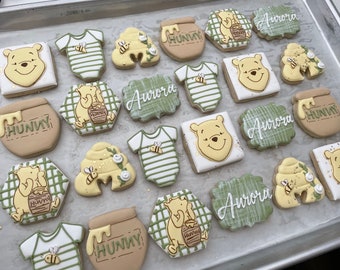 May include: A collection of Winnie the Pooh themed sugar cookies decorated with green, yellow, and brown icing. The cookies feature Winnie the Pooh, honey pots, beehives, and baby onesies. The cookies are arranged on a silver tray. The text "Hunny" and "Aurora" are written on some of the cookies.