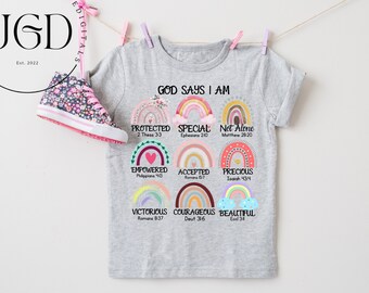 Puede incluir: Una camiseta gris con un diseño de arcoíris y el texto "Dios dice que soy" con versículos bíblicos enumerados debajo de cada arcoíris.