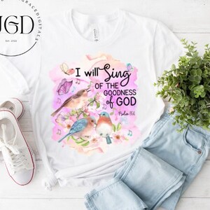 Puede incluir: Camiseta blanca con un diseño de acuarela de pájaros cantando en una rama con flores rosas. El texto "I will sing of the goodness of God Psalm 145:7" está impreso en la camiseta.