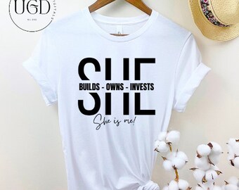Op de afbeelding: Wit t-shirt met de zwarte tekst "SHE BUILDS - OWNS - INVESTS SHE She is me!". Het shirt hangt aan een houten hanger. Op de achtergrond is een strohoed met een bloemenband te zien.
