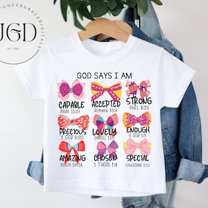 May include: White t-shirt with a colorful bow design and the text "God Says I Am" with bible verses listed below each bow.