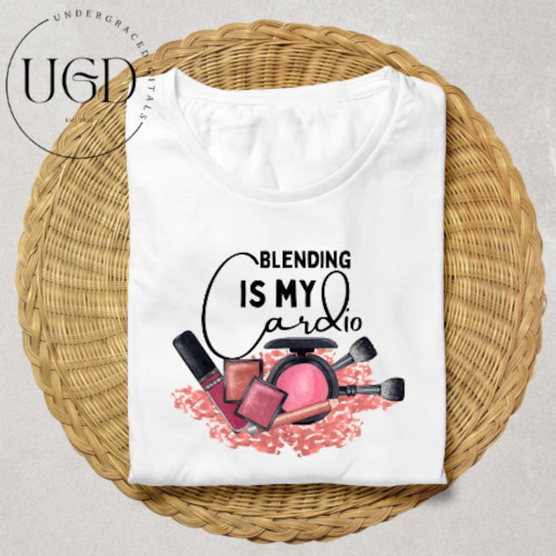 Puede incluir: Camiseta blanca con el texto "Blending is My Cardio" en escritura negra. La camiseta presenta un dise&ntilde;o de acuarela de productos de maquillaje, incluyendo l&aacute;piz labial, colorete y brochas. La camiseta est&aacute; doblada sobre una superficie circular tejida.