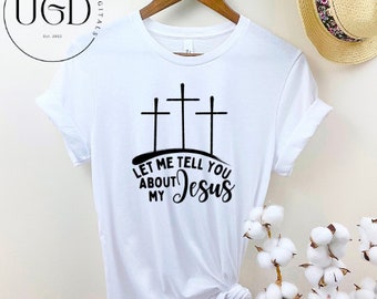 Op de afbeelding: Wit T-shirt met een zwarte afbeelding van drie kruisen en de tekst "LET ME TELL YOU ABOUT MY Jesus". Het T-shirt hangt aan een houten hanger.