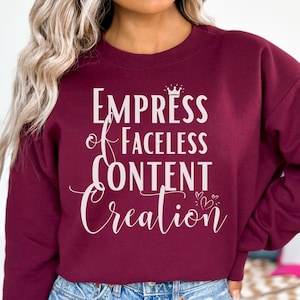 May include: A maroon sweatshirt with the text "Empress of Faceless Content Creation" in white. The text includes a crown and heart graphics. The sweatshirt is a crew neck style.