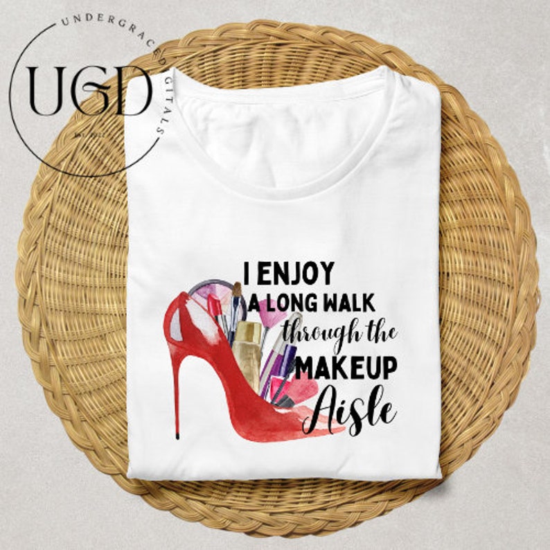 Puede incluir: Camiseta blanca con el texto &laquo;Me gusta dar un largo paseo por el pasillo de maquillaje&raquo;. El dise&ntilde;o presenta un zapato de tac&oacute;n rojo lleno de brochas de maquillaje, pintalabios y otros productos de belleza. La camiseta est&aacute; doblada sobre una superficie tejida.