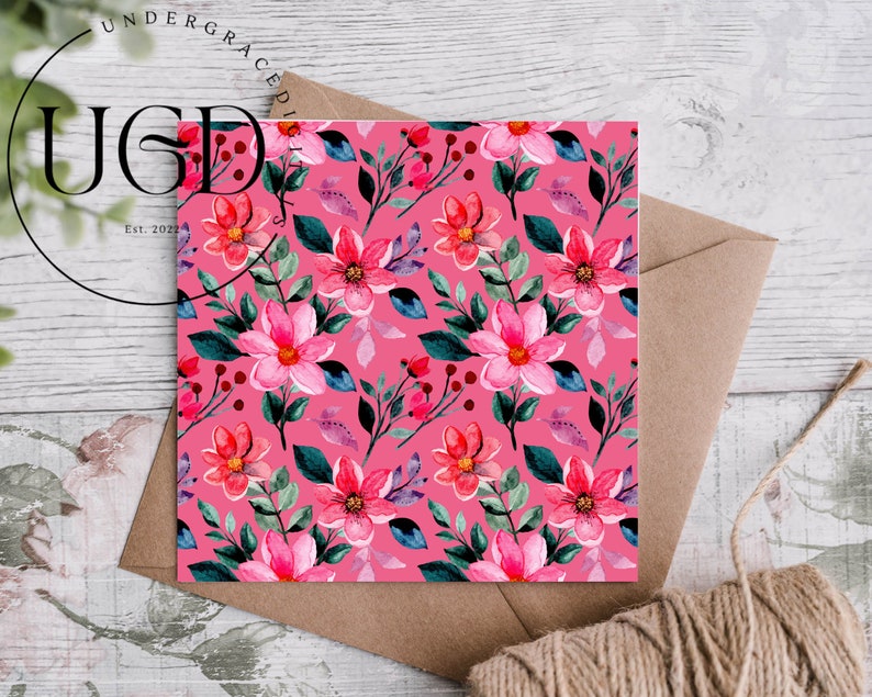 Op de afbeelding: Een vierkante kaart met een levendig roze bloemenpatroon met roze bloemen in aquarel, groene bladeren en rode bessen. De kaart ligt op een bruine envelop met een touwtje. De tekst "UNDERGRACED" en "Est. 2022" is zichtbaar.
