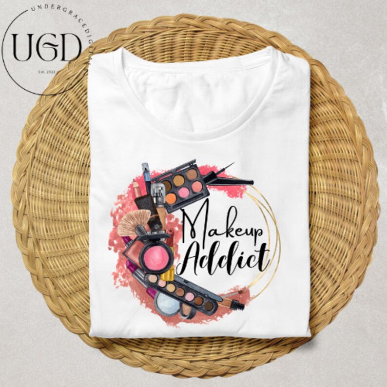 Puede incluir: Camiseta blanca con un dise&ntilde;o de acuarela de productos de maquillaje, incluyendo paletas de sombras de ojos, brochas y pintalabios, dispuestos alrededor de las palabras "Makeup Addict" en un marco dorado. La camiseta est&aacute; doblada sobre una superficie tejida.