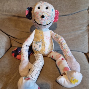 Op de afbeelding: Een patchwork-aapenspeelgoed met een wit gezicht, zwarte ogen en een roze en wit gepatroneerd lichaam. De aap zit op een beige bank.
