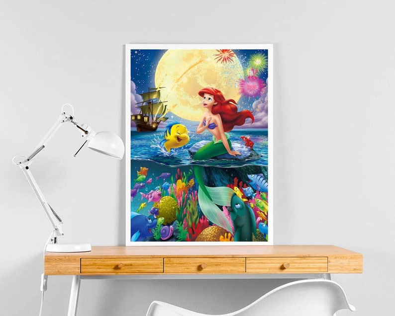 Peut inclure: Une illustration color&eacute;e d'Ariel, la Petite Sir&egrave;ne, assise sur un rocher dans l'oc&eacute;an. Elle est entour&eacute;e de poissons et de coraux. La lune est en arri&egrave;re-plan avec des feux d'artifice.