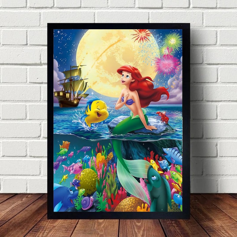 Peut inclure: Une illustration encadr&eacute;e d'Ariel, la Petite Sir&egrave;ne, assise sur un rocher avec Polochon nageant &agrave; ses c&ocirc;t&eacute;s. La sc&egrave;ne se d&eacute;roule sous l'eau avec une pleine lune, des feux d'artifice et des r&eacute;cifs coralliens color&eacute;s.
