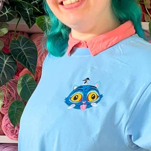 Puede incluir: Camiseta azul claro con cuello rosa con un gato de dibujos animados bordado con ojos amarillos y un sombrero azul. El fondo incluye estantes llenos de madejas de hilo coloridas y plantas verdes.