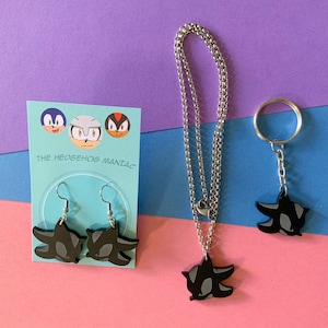 May include: A set of black Shadow the Hedgehog earrings, a necklace, and a keychain. The earrings and necklace feature a silver chain. The keychain has a silver ring and chain. The background is a multi-colored surface. The card in the image reads "The Hedgehog Maniac".