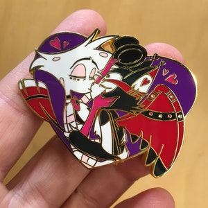Puede incluir: Un pin de esmalte dorado con dos personajes de dibujos animados, un personaje blanco y morado con un lazo rojo y un personaje negro y rojo con un sombrero de copa negro, besándose dentro de una forma de corazón. El corazón es morado con corazones rosas en la parte superior.