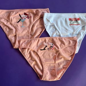 Op de afbeelding: Drie paar roze en witte ondergoed met verschillende grafische prints. Een paar heeft een cartoonfiguur met een martini-glas, een ander paar heeft een cartoonfiguur met een naamsticker waarop "Hello Loser" staat, en het derde paar heeft een cartoonfiguur met een martini-glas.