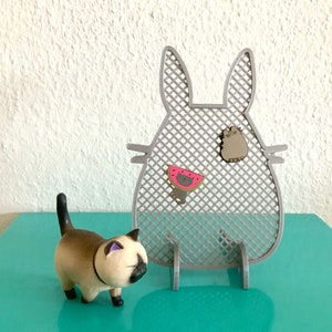 Könnte beinhalten: Ein grauer, katzenförmiger Nadelhalter mit einem Gittermuster. Der Halter hat zwei kleine Katzennadeln daran befestigt, eine mit einer Wassermelone und die andere mit einem einfachen Katzendesign. Eine kleine, hellbraune Katzenfigur befindet sich im Vordergrund.