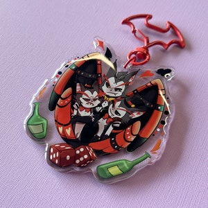 Op de afbeelding: Een transparante acryl sleutelhanger met drie cartoonfiguren met rode, zwarte en witte details. De figuren zijn omringd door rode harten, dobbelstenen en groene flessen. Een rode, vleermuisvormige metalen hanger is aan de bovenkant van de sleutelhanger bevestigd.